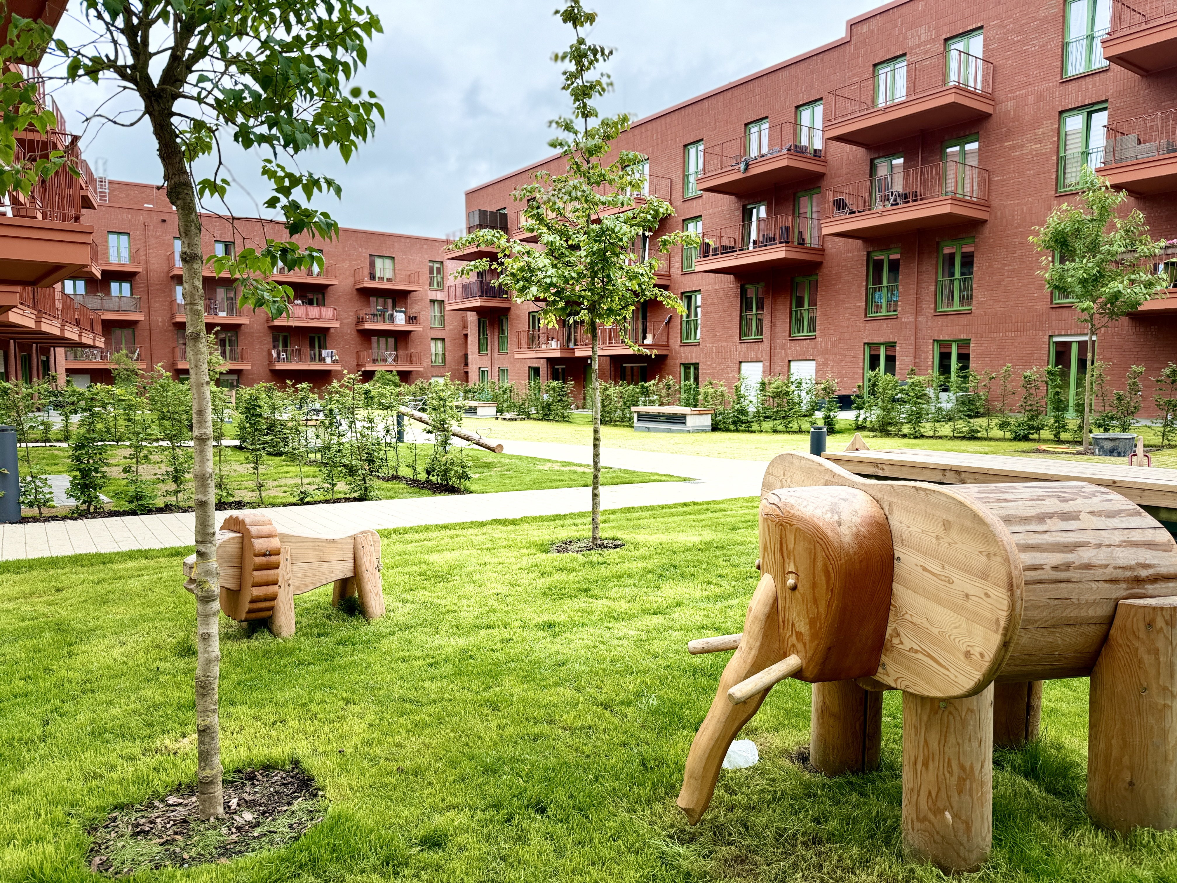Holzskulptur eines Elefanten auf einer Rasenfläche vor einem roten Wohngebäude mit Balkonen und jungen Bäumen