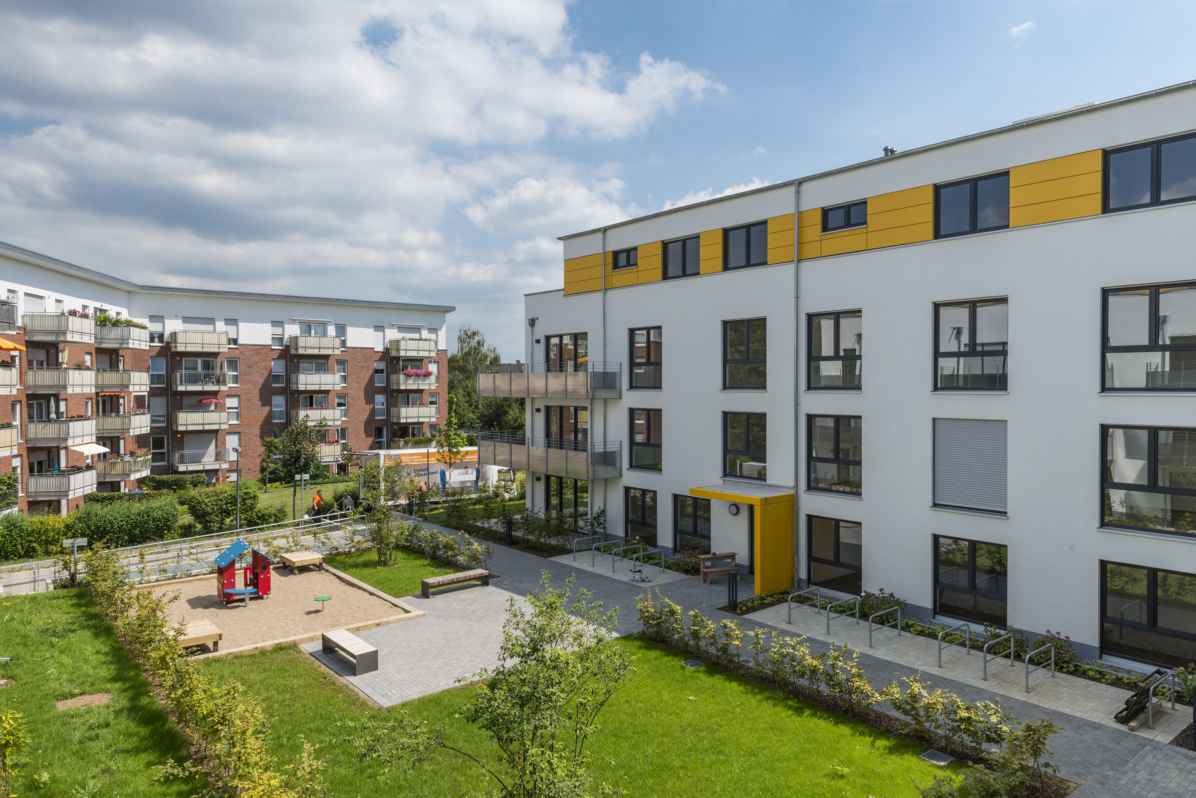 Mehrstöckiges modernes Wohngebäude mit gelben Akzenten neben einem Spielplatz und angrenzenden weiteren Wohnhäusern unter bewölktem Himmel