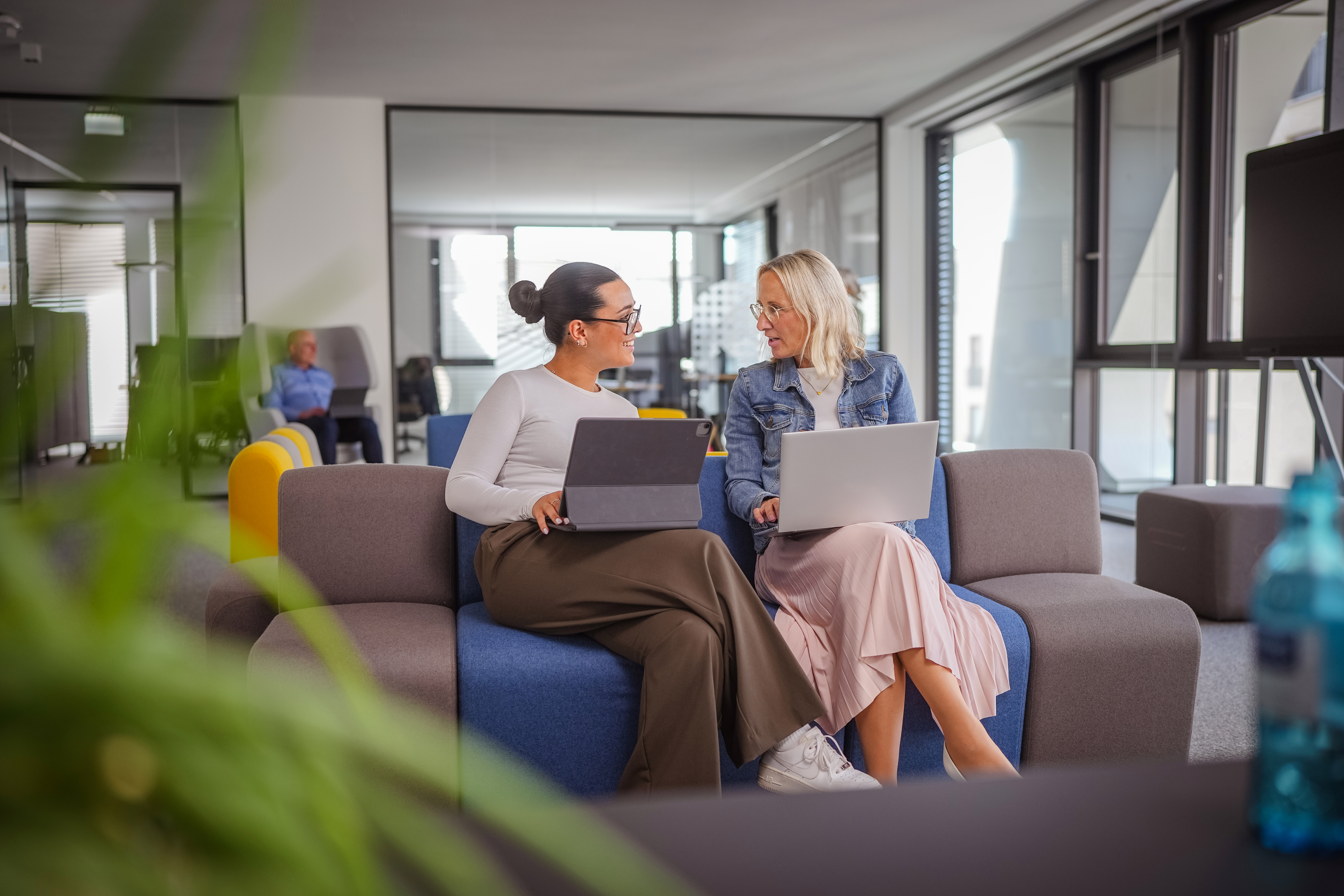 Zwei Frauen sitzen auf einem Sofa im Büro und unterhalten sich mit geöffneten Laptops auf dem Schoß